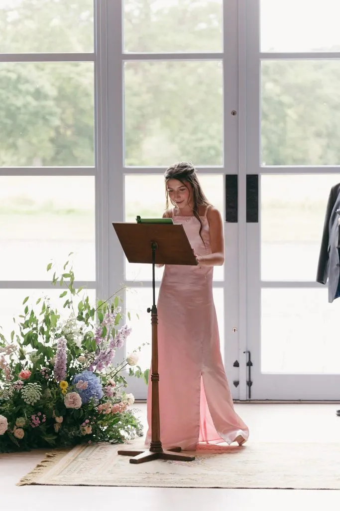 fraoch floral design woman in a pale pink dress standing at a wooden lectern, reading from a tablet, with a large glass-windowed backdrop and floral arrangements nearby.
