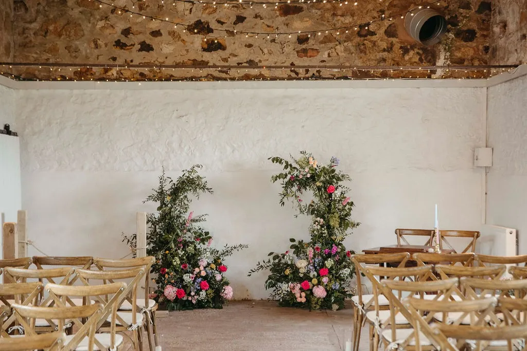 fraoch floral design rustic ceremony space with two flower arches, string lights, and wooden chairs in a whitewashed room.