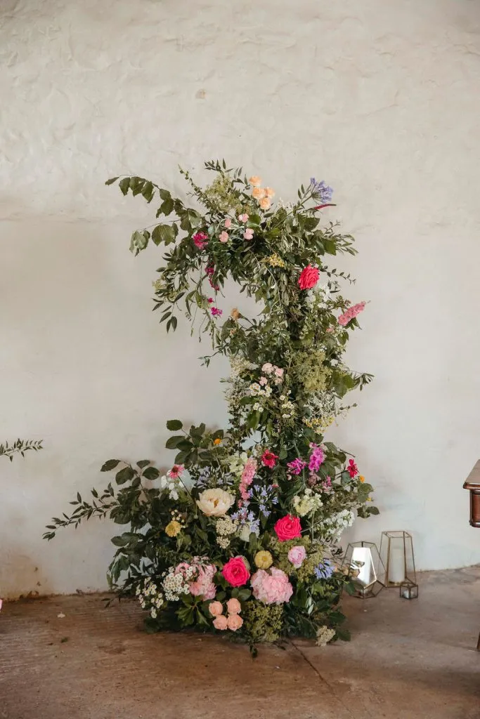 fraoch floral design tall standing floral arrangement with pink, peach, and white roses and green foliage against a plain beige wall.
