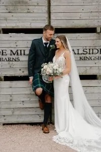 fraoch floral design bride and groom pose together against a weathered wooden wall; she holds a bouquet of white and blush flowers, he wears a dark formal jacket with a green tartan kilt and brown boots, smiling at each other under her veil.