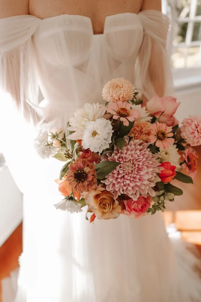 fraoch floral design bride holding a pastel bouquet of pink and white flowers against her off-the-shoulder wedding dress.