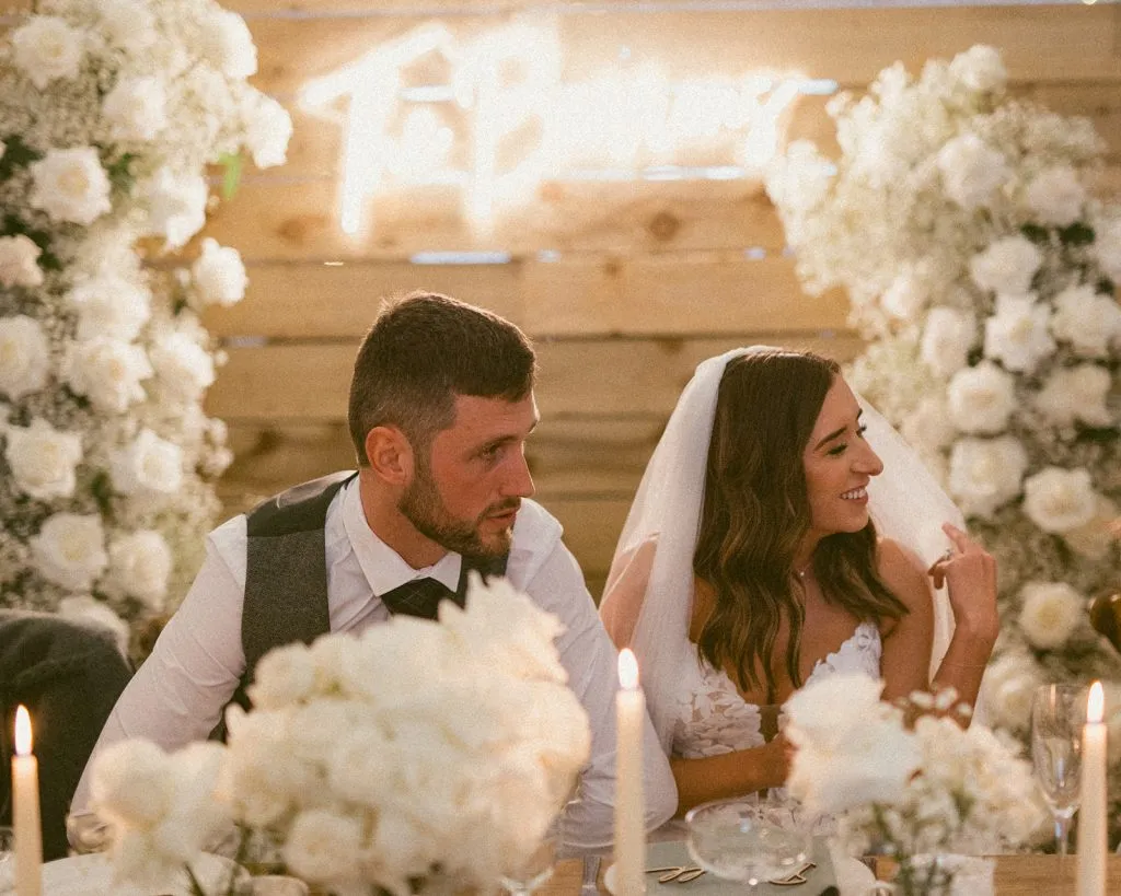 fraoch floral design bride and groom sit at a candlelit wedding reception table flanked by white flowers, smiling at each other.