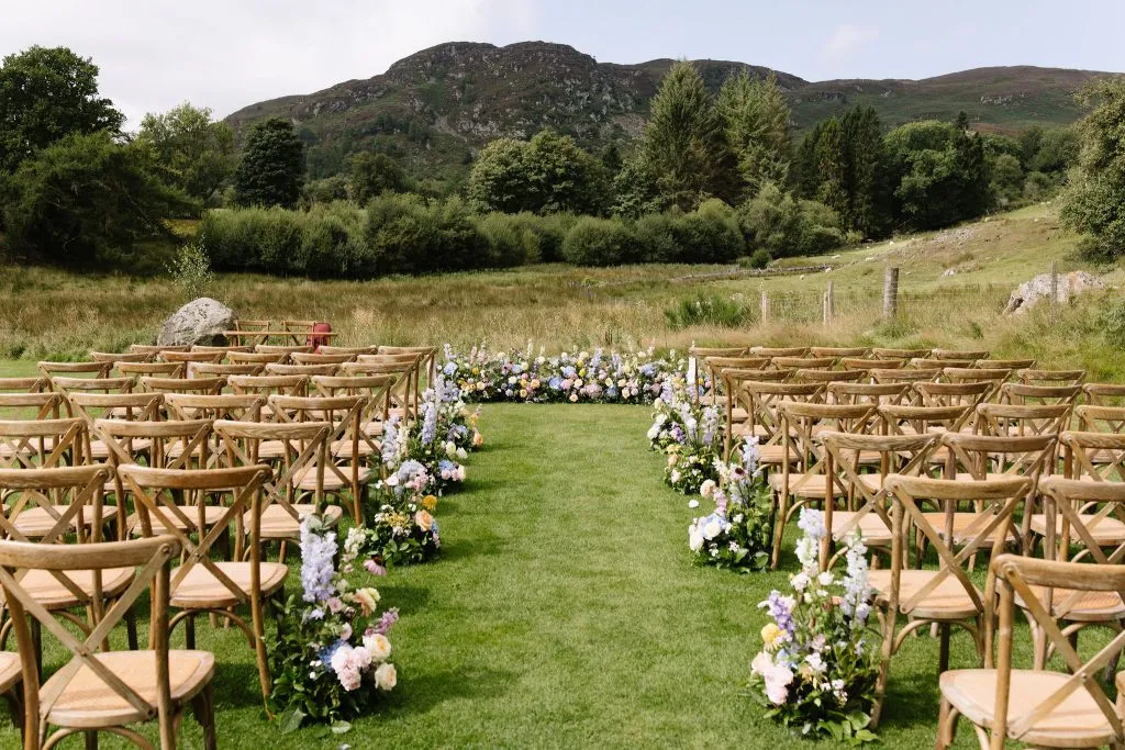 fraoch floral design outdoor wedding ceremony setup with wooden chairs arranged in rows along a grassy aisle, bordered by colorful flower arrangements and a rural landscape with mountains in the background.