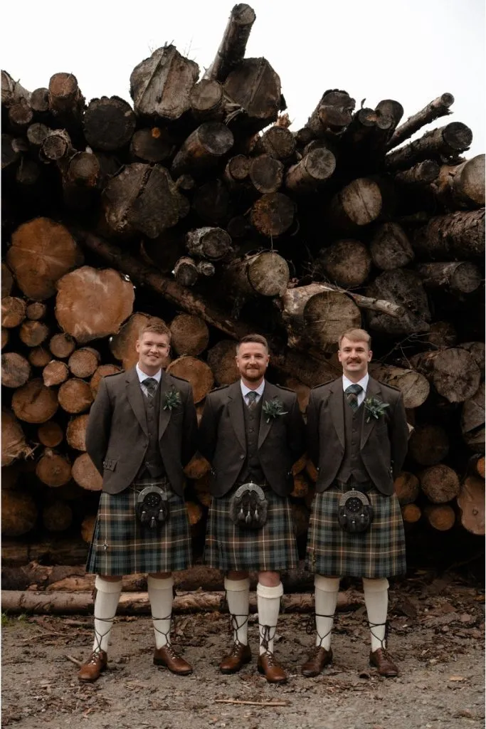 fraoch floral design three men in matching brown suits and green plaid kilts stand in front of a stacked log pile, smiling for a wedding photo.