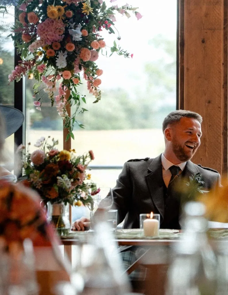 fraoch floral design man in a suit at a wedding reception, smiling, seated at a table with colorful flower arrangements and a lit candle nearby.