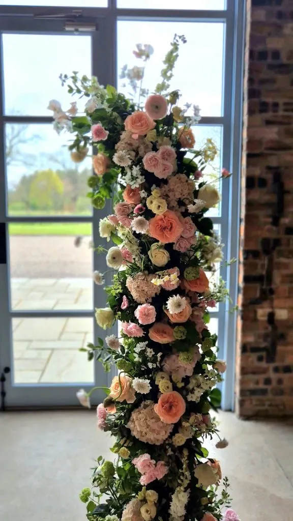 fraoch floral design tall vertical floral arrangement of pastel pink, peach, and white roses with white blossoms and greenery, set indoors by a large window.