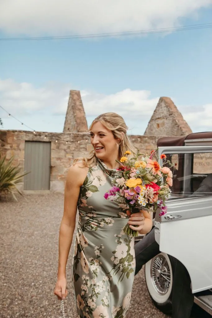 fraoch floral design woman in a light green floral dress holding a colorful bouquet, standing beside a vintage car outdoors.