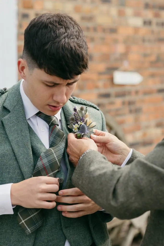 fraoch floral design young man in a green tweed suit watches as a boutonniere is pinned to his lapel by another person outdoors.