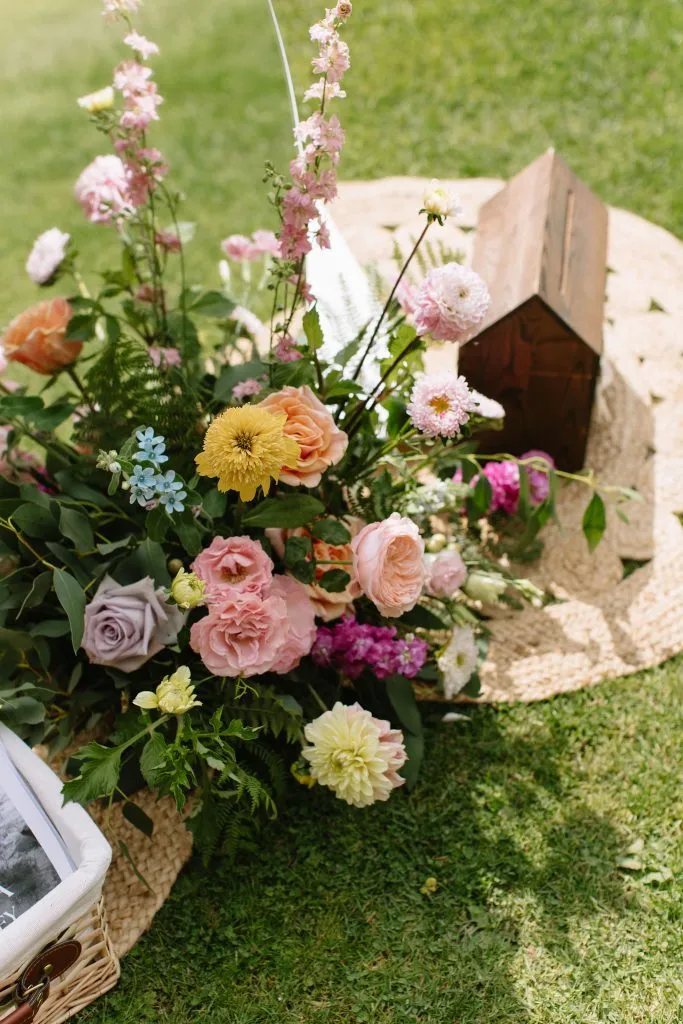 fraoch floral design colorful outdoor floral arrangement with pink, peach, and yellow blooms in a wicker basket on grass, with a small wooden birdhouse in the background