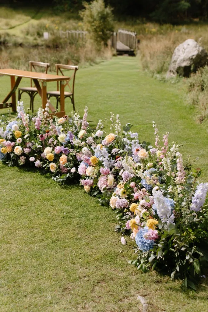 fraoch floral design colorful flower arrangement along a lawn with a wooden table and chairs on the left, and a grassy path leading to a small bridge in the distance.