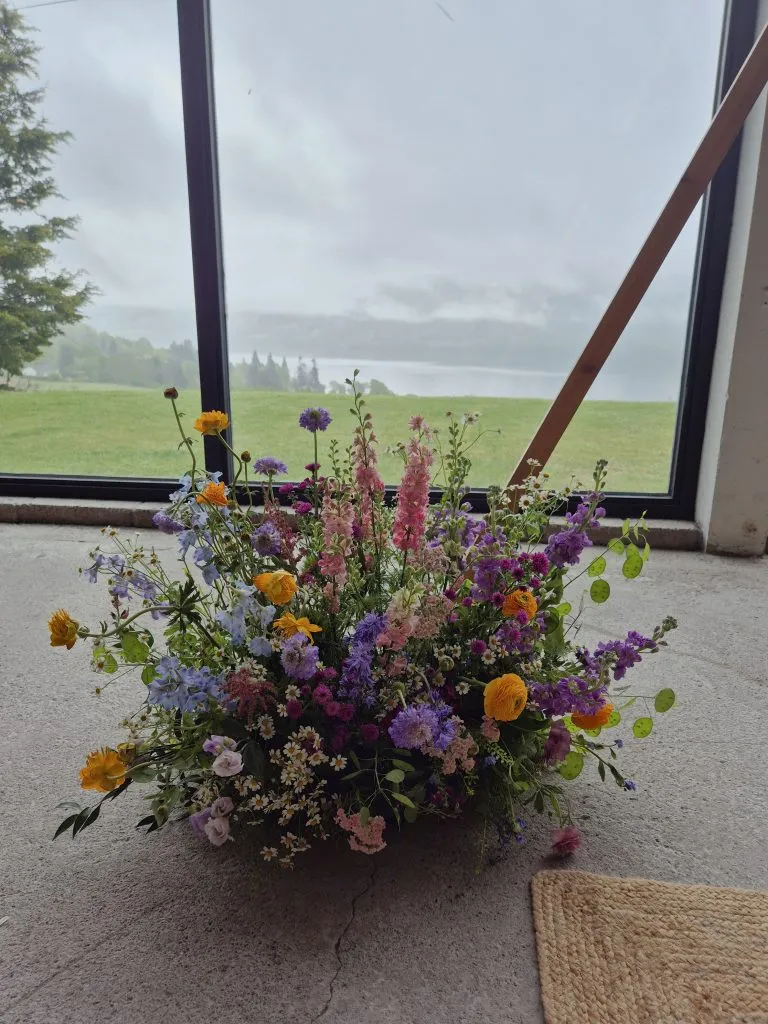 fraoch floral design a large colourful arrangement of wildflowers sits on a concrete floor by a slanted window, with a grassy landscape outside.