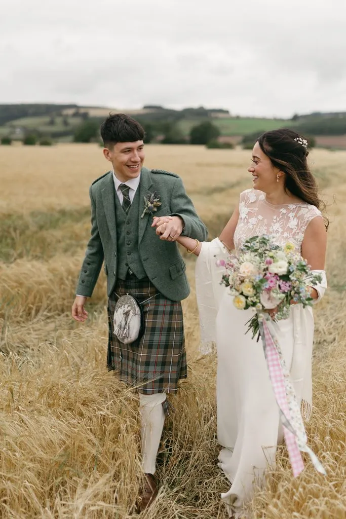 fraoch floral design bride and groom walk hand-in-hand through a golden field; she carries a floral bouquet in a lace dress, he wears a green tweed jacket and kilt.