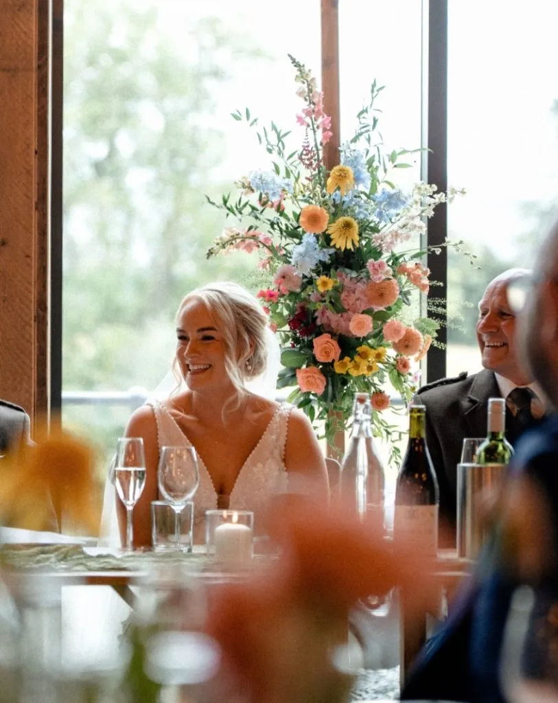 fraoch floral design bride smiling at a wedding reception, seated at a table with a tall colorful floral centerpiece, champagne glasses and candles beside her.