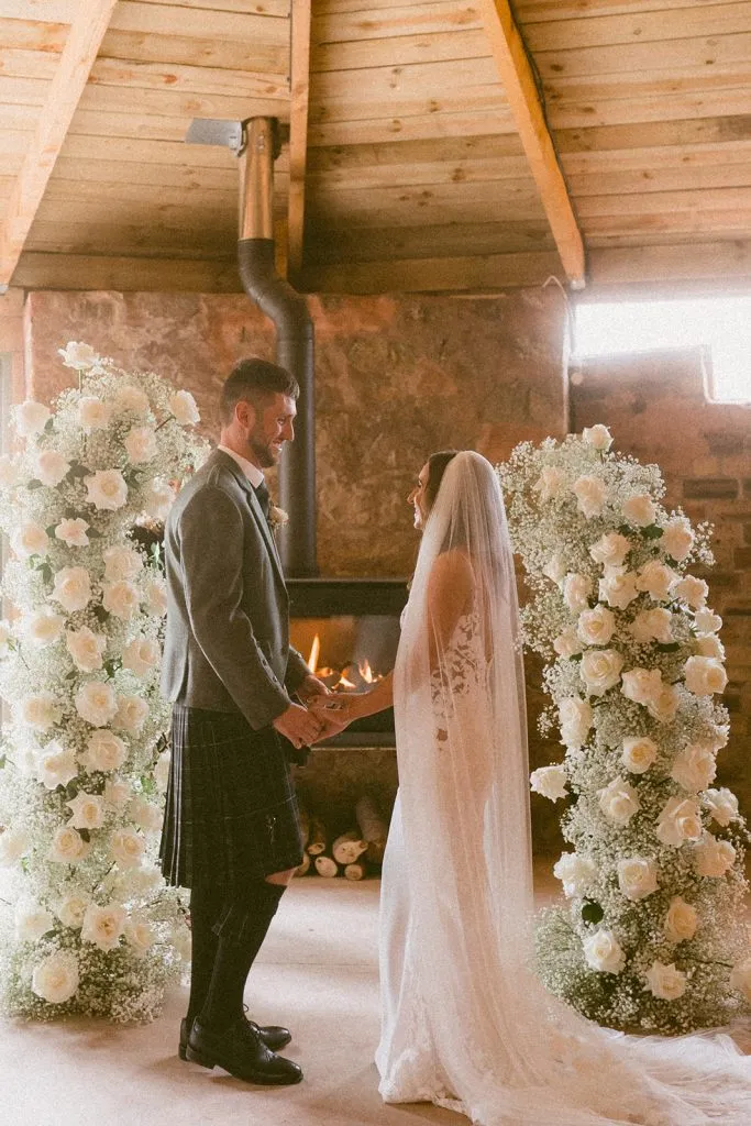 fraoch floral design bride and groom hold hands facing each other between tall white rose arches in a rustic indoor wedding by a fireplace.