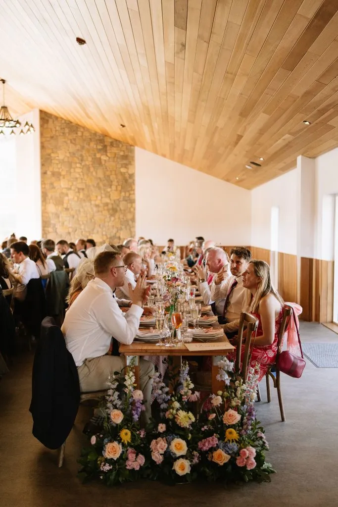 fraoch floral design guests at a long banquet table in a rustic hall with a wooden ceiling and stone wall, wedding floral arrangements in the foreground