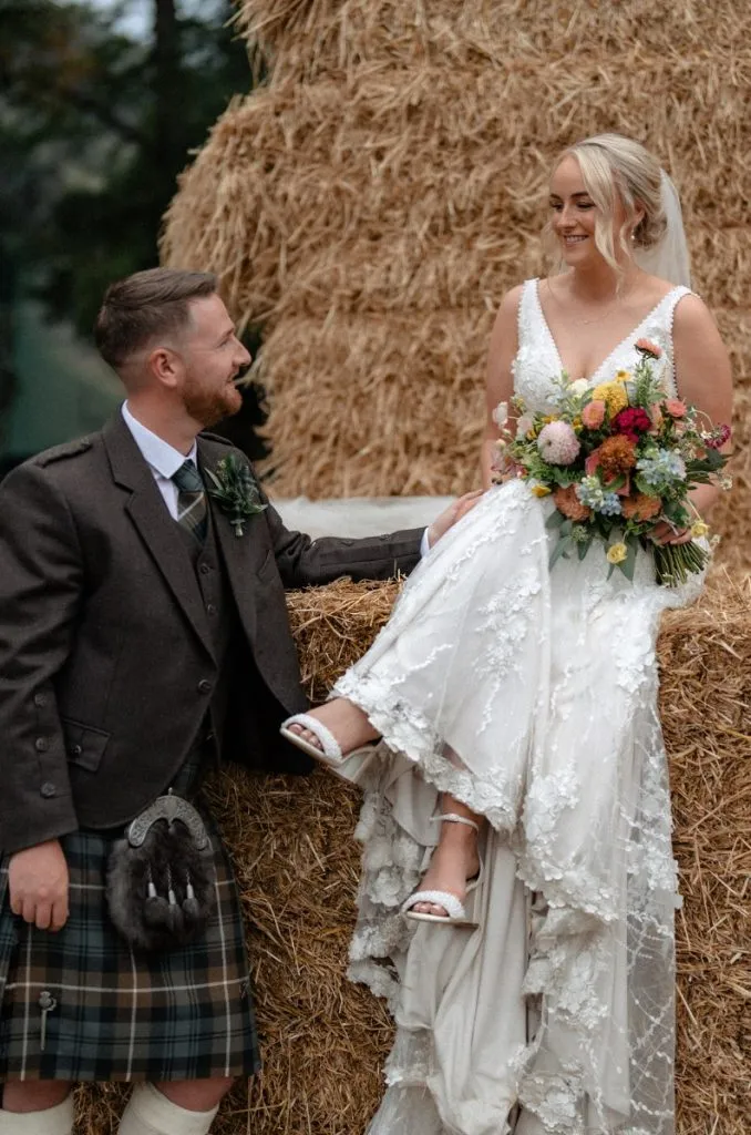 fraoch floral design bride in a lace wedding gown sits on a hay bale while a groom in a tartan kilt looks at her, them smiling together in a rustic outdoor setting.