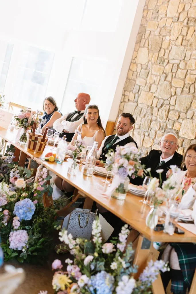 fraoch floral design wedding guests in formal attire sit along a long wooden table adorned with pastel flowers in a bright room with a stone wall behind them.