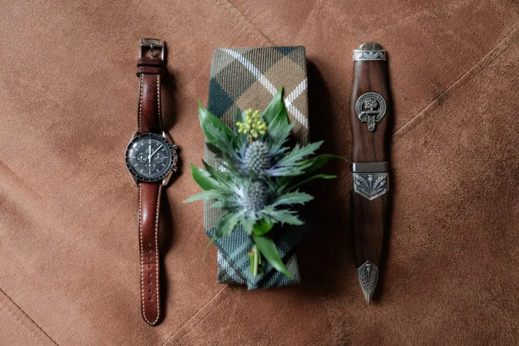 fraoch floral design left: a brown leather-strap wristwatch, center: a plaid-wrapped gift with greenery, right: a decorative carved wooden dagger on a brown suede surface