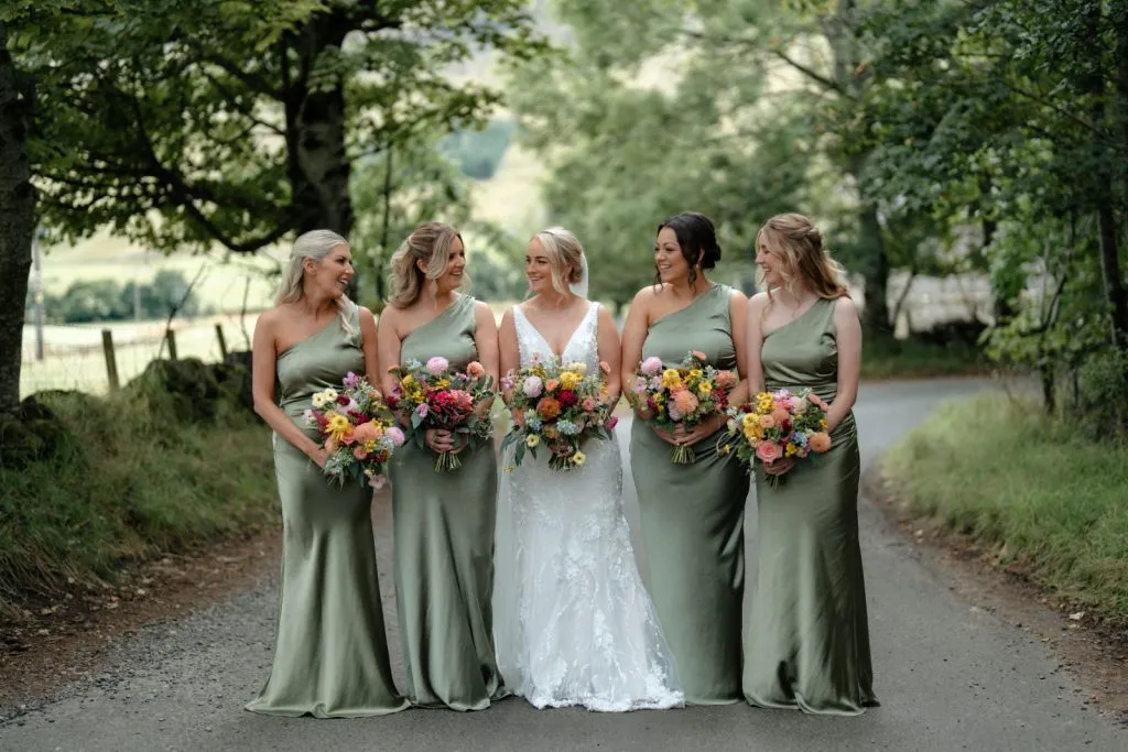 fraoch floral design bride in a white lace gown walking with four bridesmaids in sage green dresses, each holding colorful bouquets on a country road.