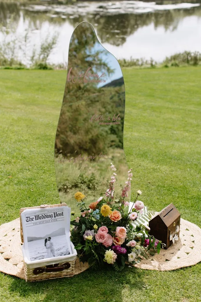fraoch floral design curved reflective wedding sign with pink text on a grassy lawn, surrounded by flowers.