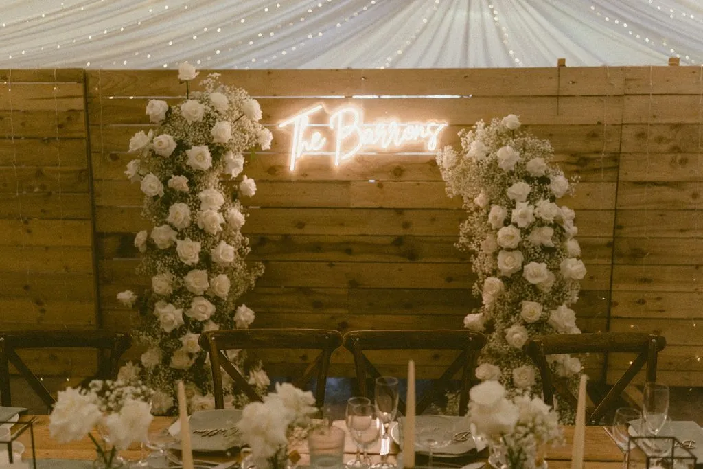 fraoch floral design wooden barn-style reception with two tall white-flower arches framing a neon'The Barn' sign above a long dining table