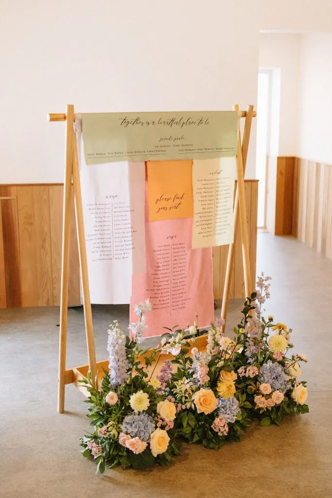 fraoch floral design wedding seating chart displayed on a wooden easel with pastel fabric banners and a flower arrangement at the base.
