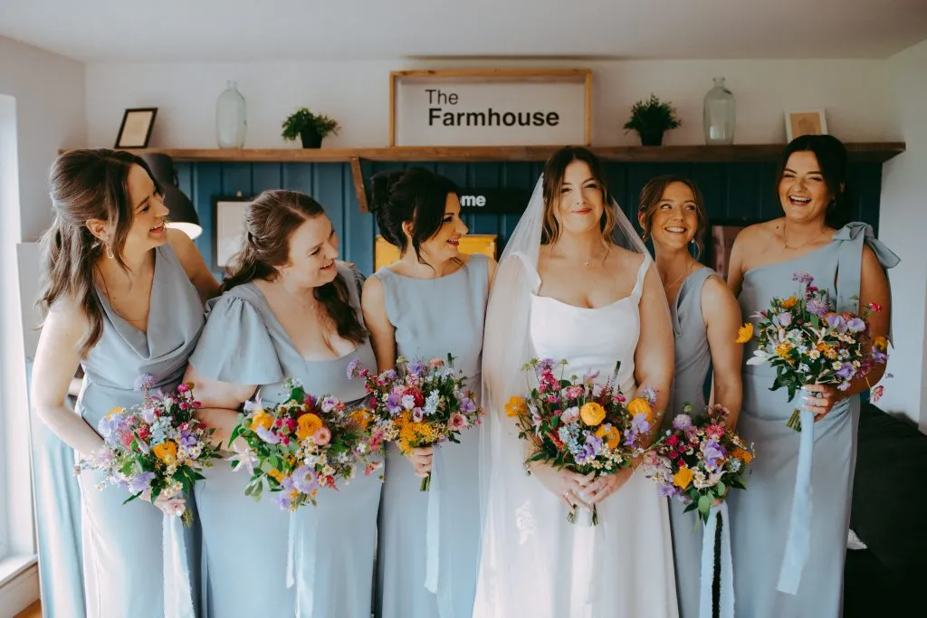 fraoch floral design bride in a white dress surrounded by bridesmaids in pale blue gowns, holding colorful bouquets indoors.