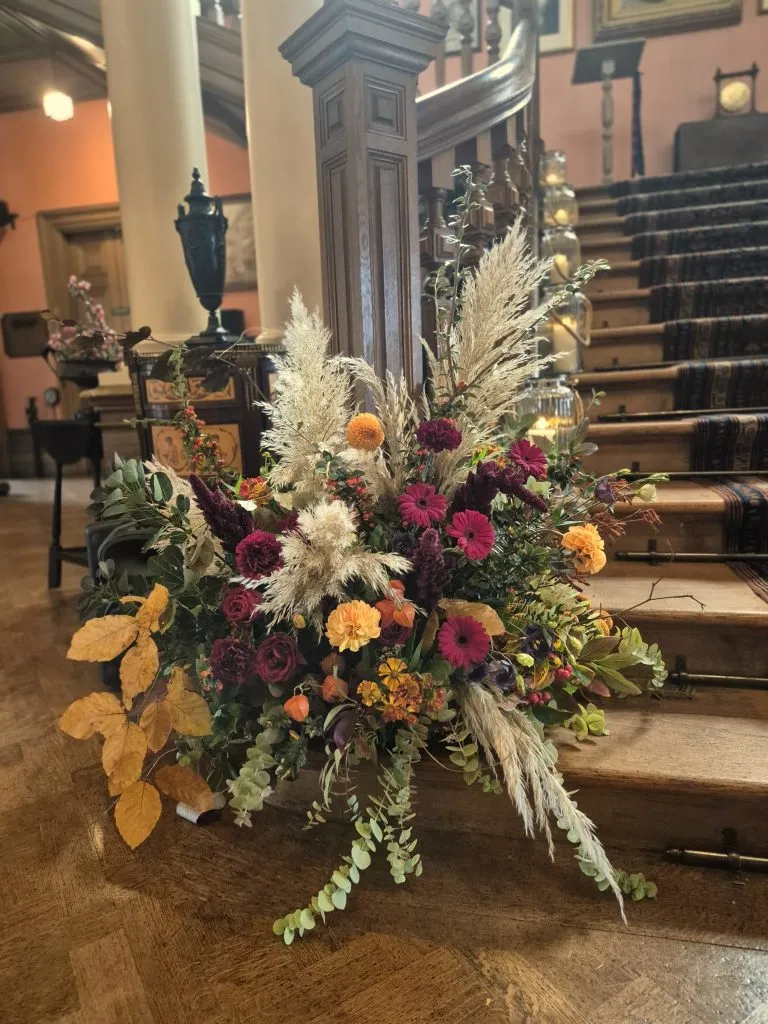 fraoch floral design autumn floral arrangement with dried grasses and colorful flowers at the base of a carved wooden staircase in a warmly lit interior.