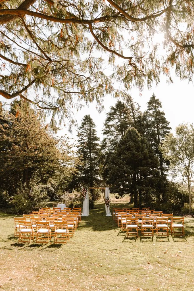 fraoch floral design outdoor wedding ceremony setup on a grassy lawn with orange wooden chairs facing a white fabric-draped arch under tall trees in sunlight.