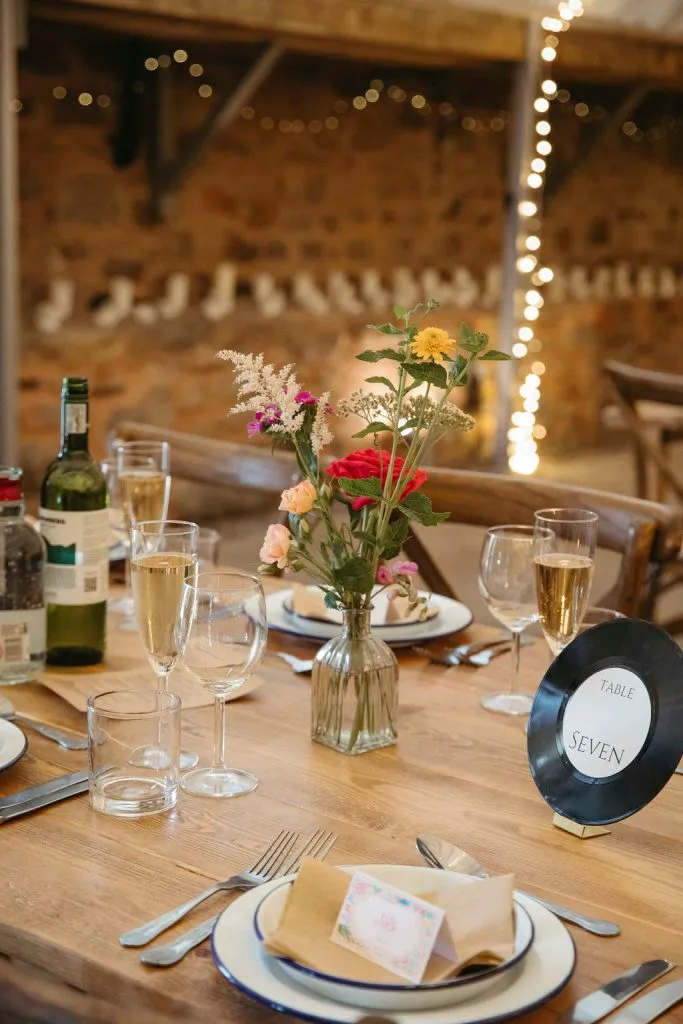 fraoch floral design table set for a formal dinner with a glass bottle, wine glasses, and a vase of mixed flowers as a centerpiece; a round table marker reads 'table seven' in the foreground.