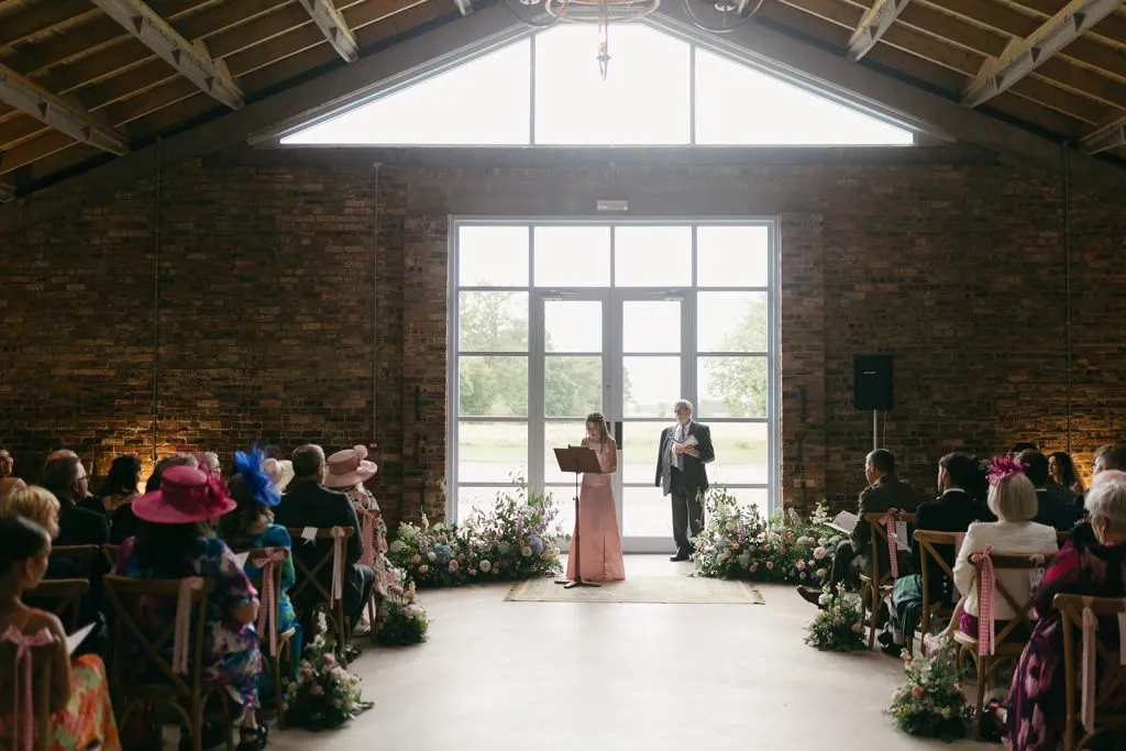 fraoch floral design bride and officiant stand at the front of a rustic brick-walled venue, reading vows as guests watch from chairs lined with floral arrangements.