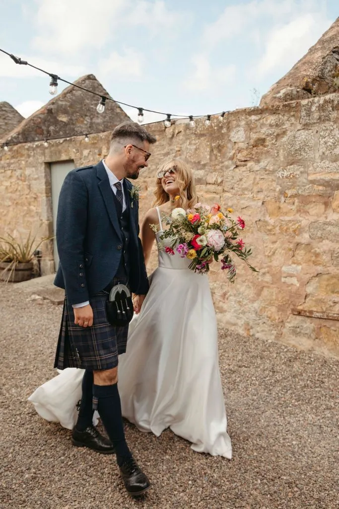 fraoch floral design a bride in a white wedding gown and a man in a kilt share a smile while holding a vibrant bouquet outdoors against a stone wall and string lights.
