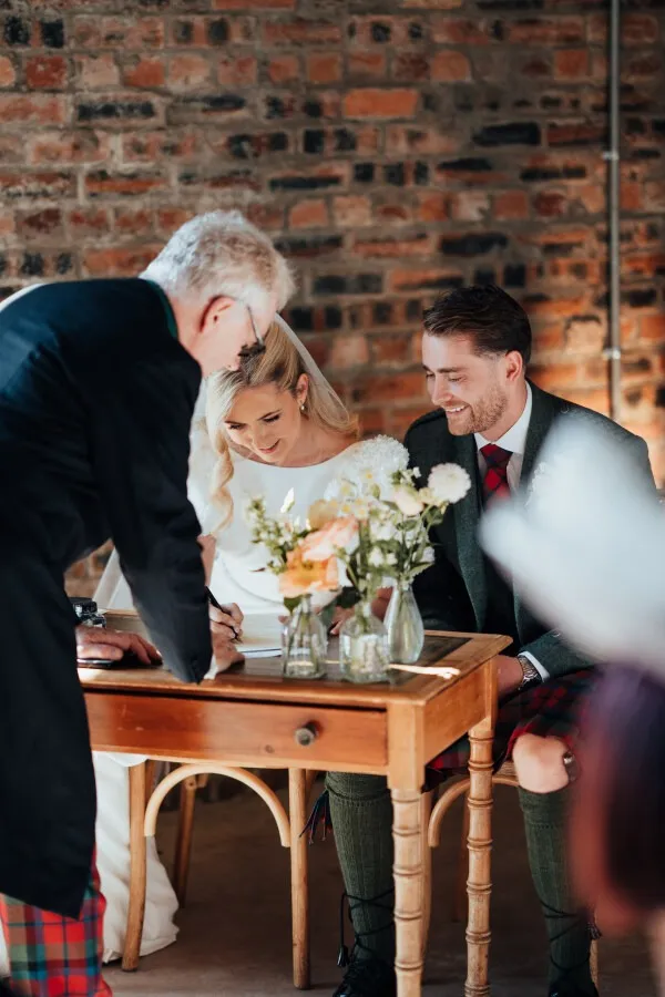 fraoch floral design bride and groom signing a document at a wooden table during their wedding, assisted by an older man, with a bouquet in front and a brick wall backdrop.