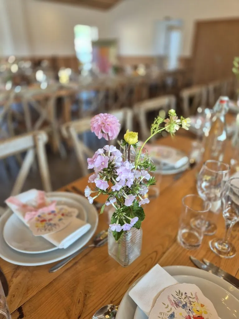 fraoch floral design pink and yellow flowers in a small glass vase on a wooden dining table, with plates, napkins, and glassware set for a meal in the foreground.