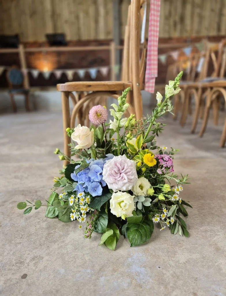 fraoch floral design a colorful flower bouquet on the floor in front of wooden chairs at an event space, with pastel accents and greenery in focus.