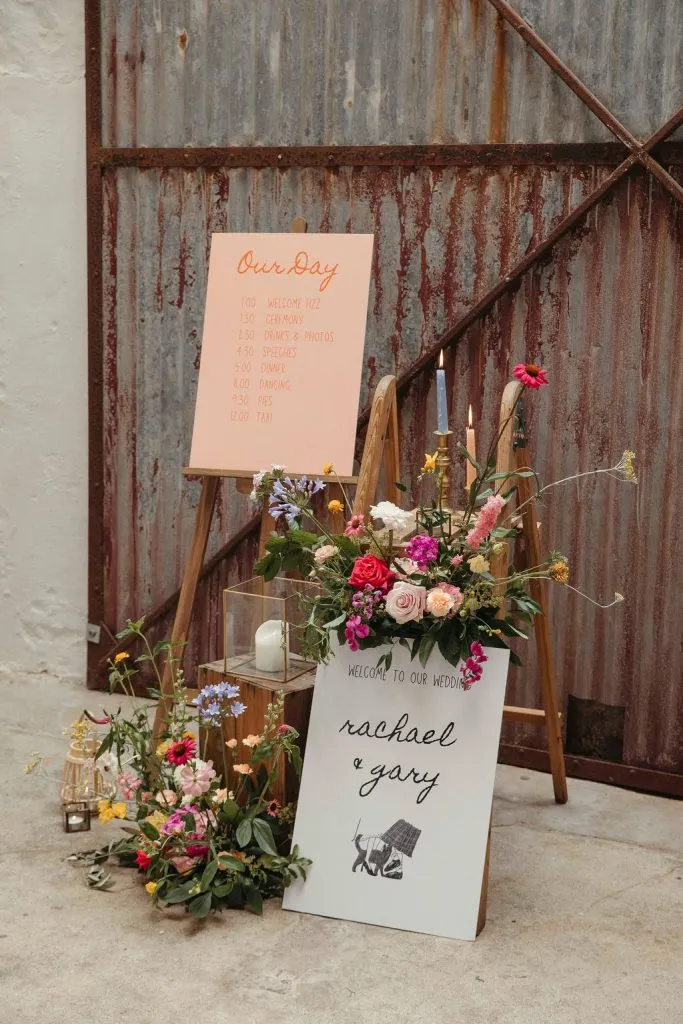 fraoch floral design rustic wedding display with a pink schedule board, colorful flowers, and a chalkboard sign reading 'rachel & gary' beside a metallic corrugated backdrop.