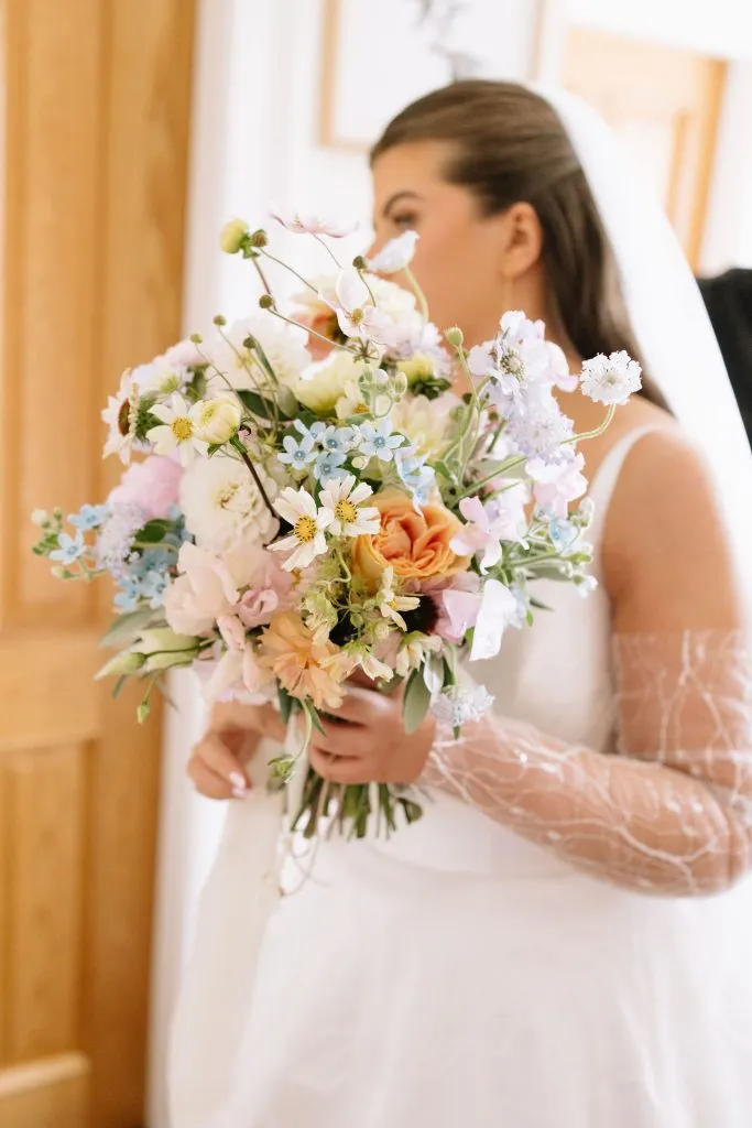 fraoch floral design bride in a white wedding dress holding a colorful pastel bouquet indoors, veil visible in the background.
