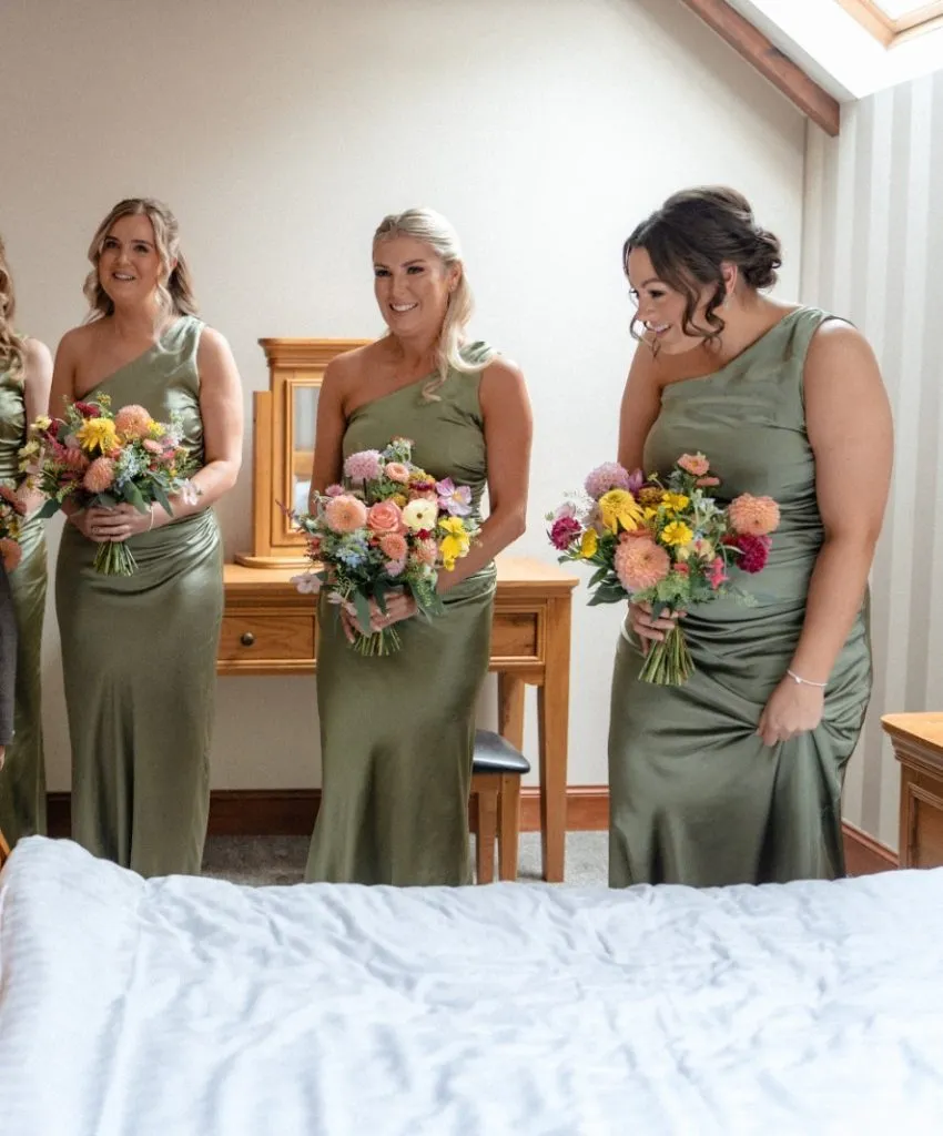 fraoch floral design bridesmaids in sage green dresses hold colorful bouquets in a bridal-suite setting, smiling together.