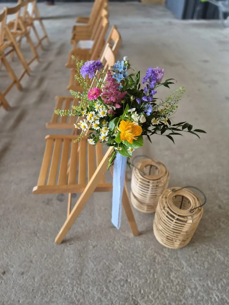 fraoch floral design colorful bouquet of wildflowers with daisies and purple blooms tied with a blue ribbon resting on a row of wooden chairs at a ceremony venue.