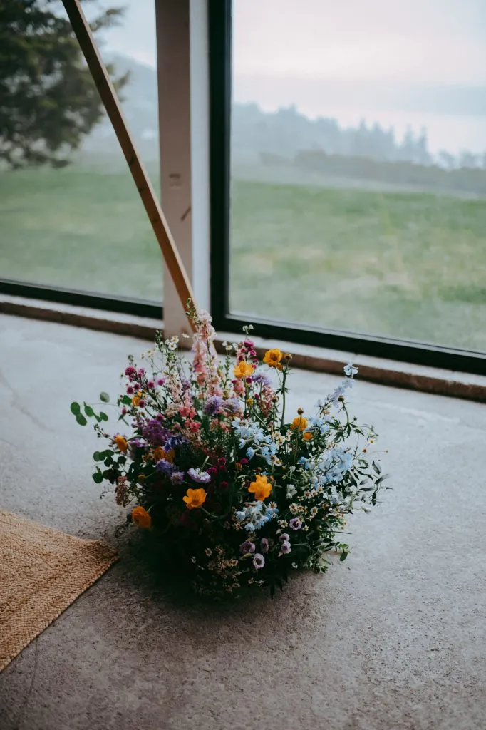 fraoch floral design bouquet of colorful wildflowers on a concrete floor by a window indoors, with a hazy green landscape outside.
