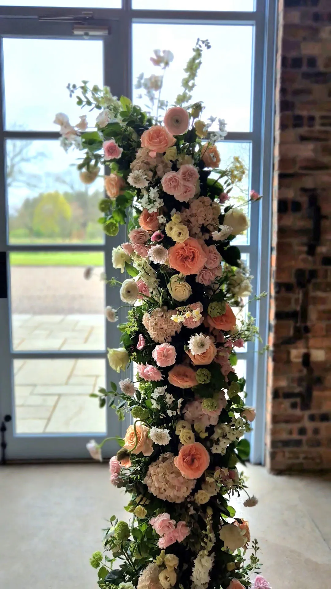 fraoch floral design tall vertical floral arrangement of pastel pink, peach, and white roses with white blossoms and greenery, set indoors by a large window.