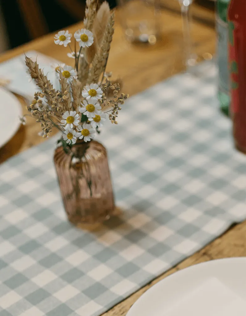 fraoch floral design vase with white daisies and dried wheat on a blue-and-white checkered tablecloth, blurred background.