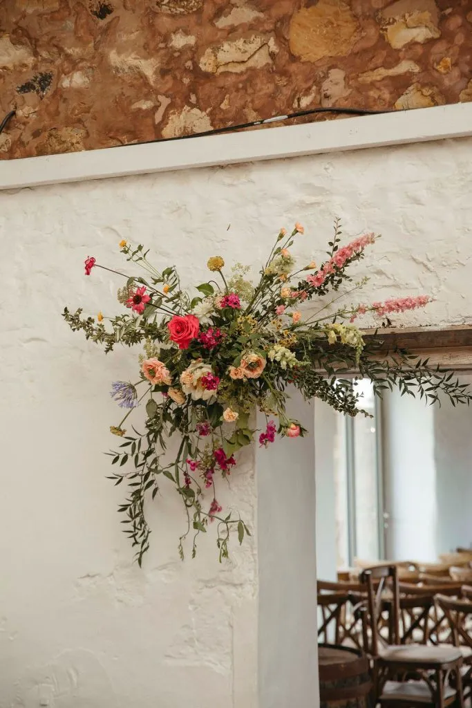 fraoch floral design decorative cascading floral arrangement mounted to a white wall with pink, peach, and yellow blooms and green foliage in a rustic interior.