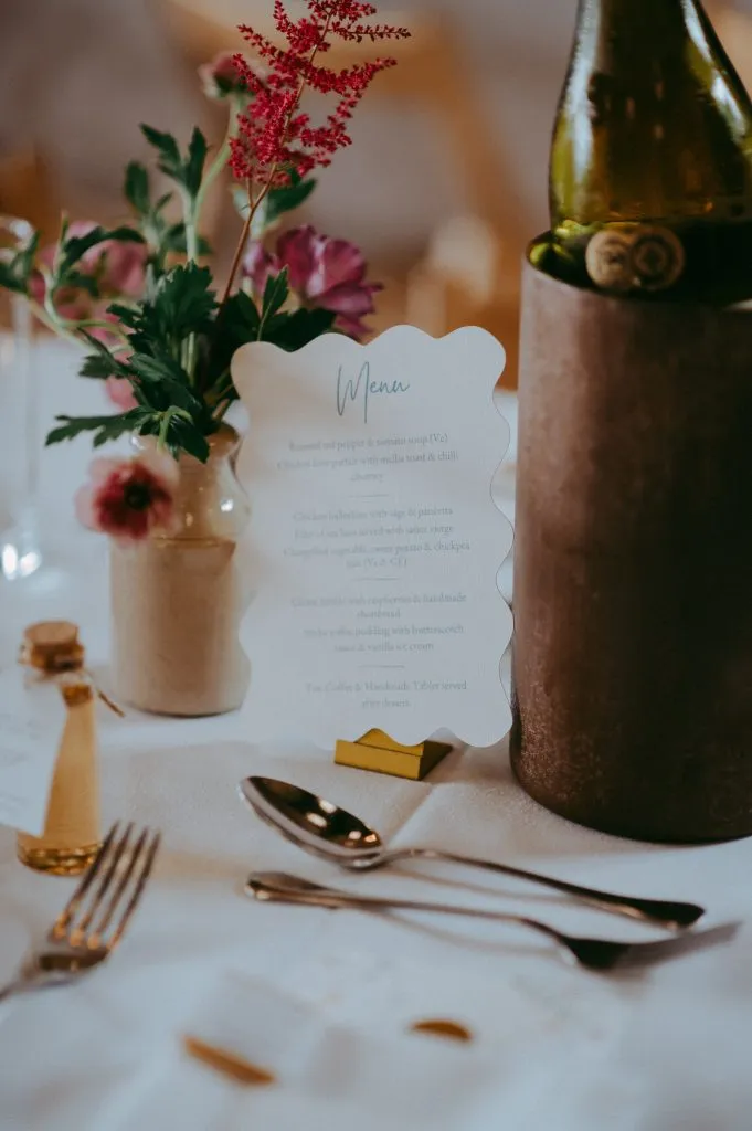 fraoch floral design elegant table setting with a floral centerpiece, a white menu card, and a wine bottle in a metal cooler on a white tablecloth.
