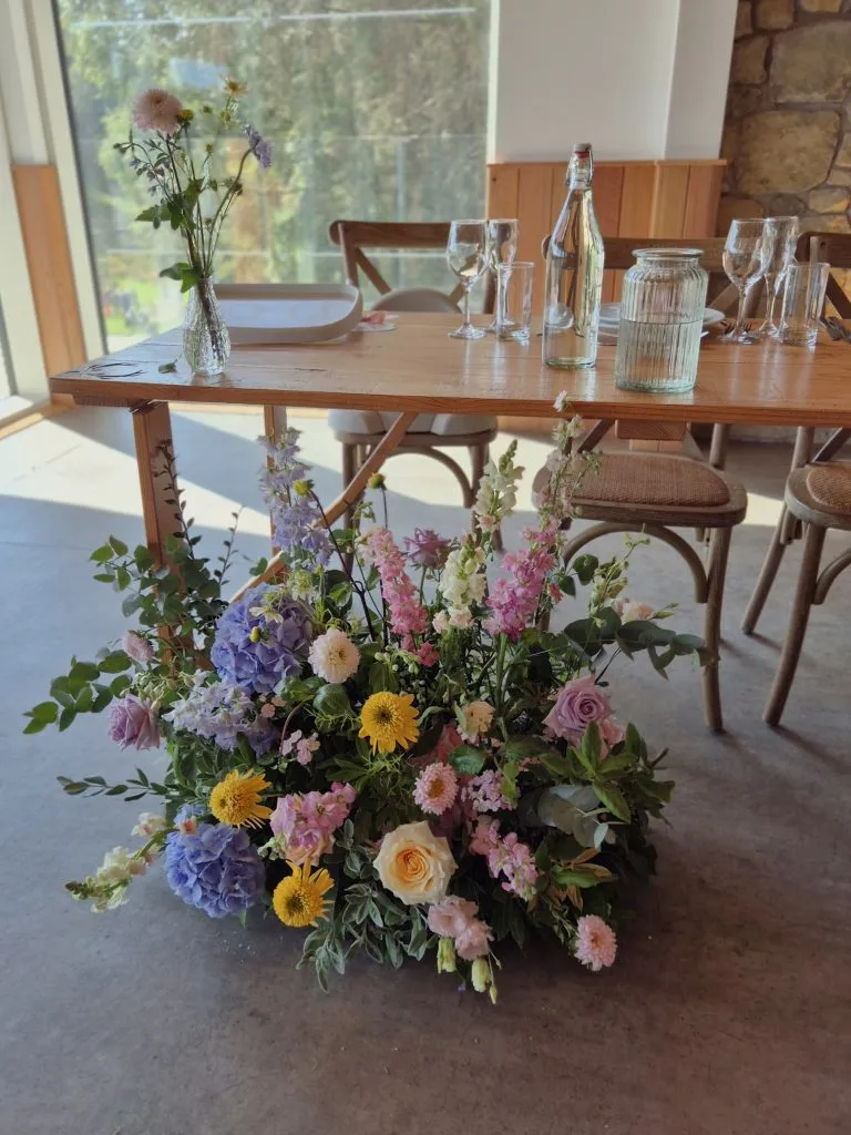 fraoch floral design wood dining table set for a meal with glassware, a bottle, and a vase of pink, purple, and yellow flowers; a large flower arrangement sits in the foreground.