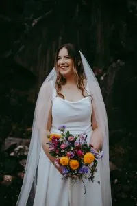 fraoch floral design bride in a white wedding dress and veil, smiling while holding a colorful bouquet outdoors.