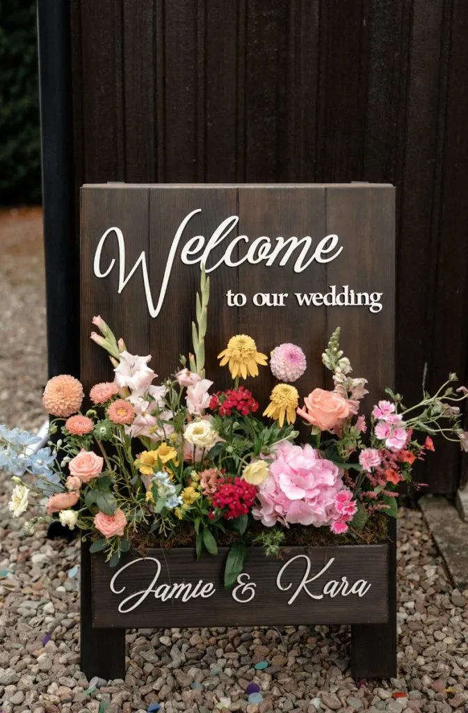 fraoch floral design wooden wedding sign reading 'welcome to our wedding' with a colorful flower arrangement; names jamie & kara at the bottom.