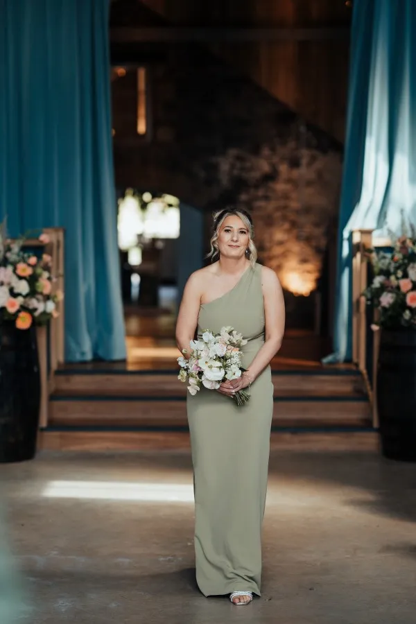 fraoch floral design a woman in a sage green one-shoulder gown holds a bouquet, standing in a decorated wedding venue with blue drapes and warm lighting.