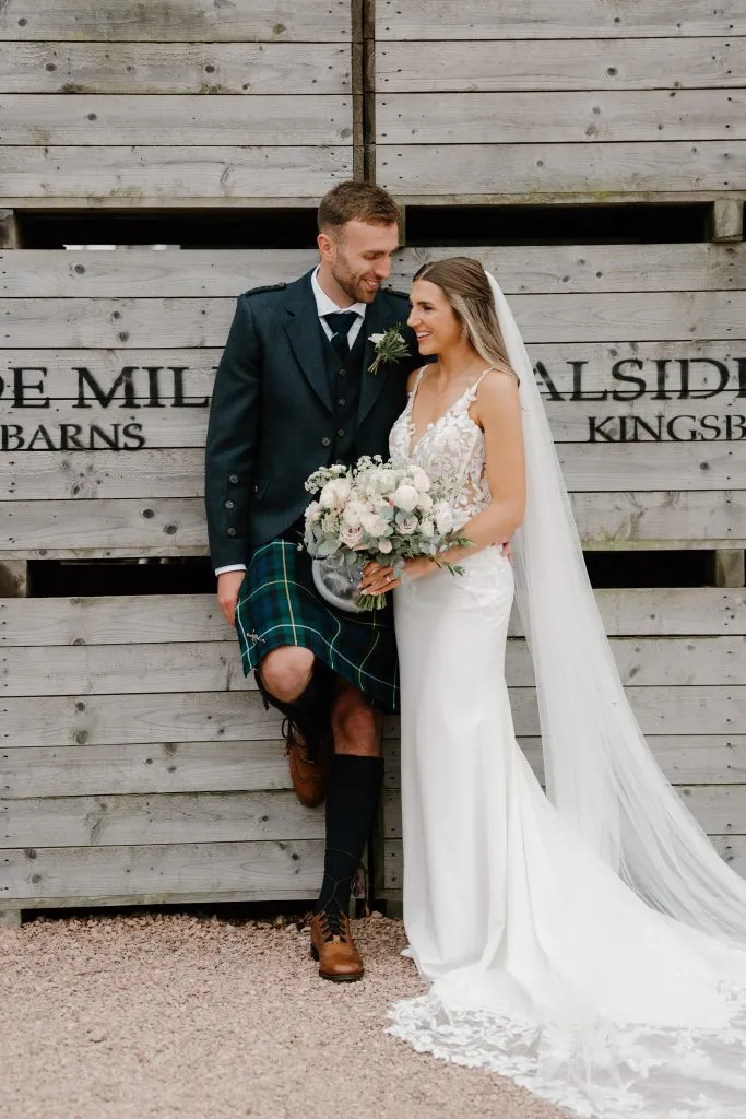 fraoch floral design bride and groom pose together against a weathered wooden wall; she holds a bouquet of white and blush flowers, he wears a dark formal jacket with a green tartan kilt and brown boots, smiling at each other under her veil.