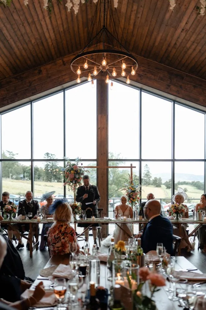 fraoch floral design man giving a speech at a wedding reception in a rustic wooden hall with a chandelier and floral centerpieces at long tables